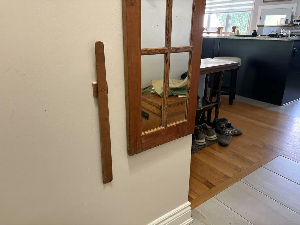 Schoenlepel van licht eiken aan een lichte muur naast een oude spiegel met houten lijst, met doorkijk naar de keuken