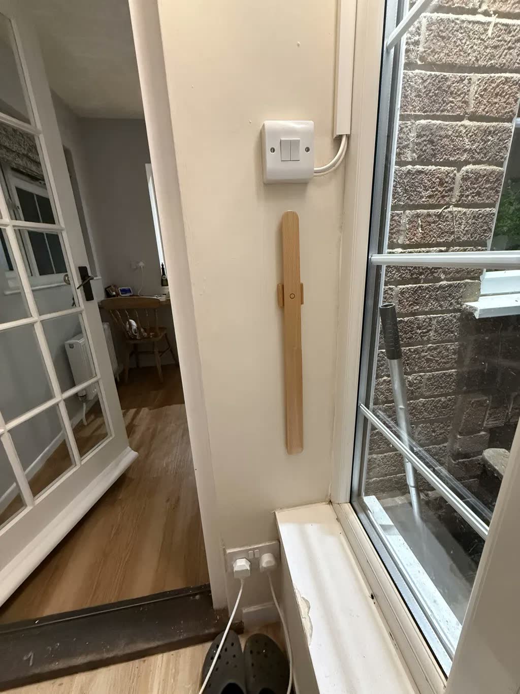 Schoenlepel van licht eiken aan een lichte muur bij een raam en deuropening, met een lichtschakelaar erboven en klompen op de vloer